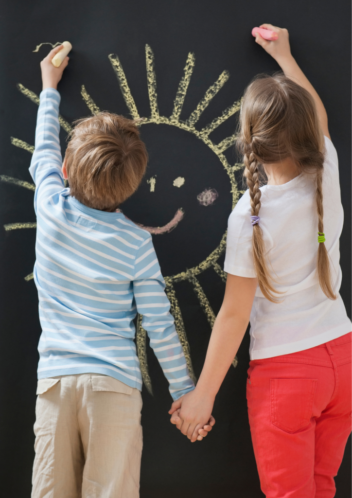 2 enfants de dos se tiennent la main et dessinent au tableau un soleil souriant
